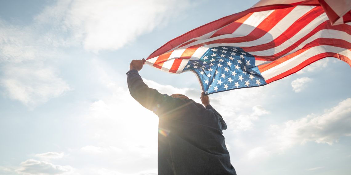 Portrait of cheerful young man looking forward while standing proudly with holding USA flag over his head against wind and blue sky, happy american holding the United states flag celebration