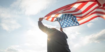 Portrait of cheerful young man looking forward while standing proudly with holding USA flag over his head against wind and blue sky, happy american holding the United states flag celebration