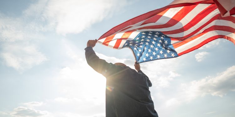 Portrait of cheerful young man looking forward while standing proudly with holding USA flag over his head against wind and blue sky, happy american holding the United states flag celebration