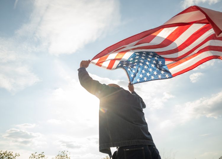 Portrait of cheerful young man looking forward while standing proudly with holding USA flag over his head against wind and blue sky, happy american holding the United states flag celebration