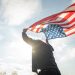 Portrait of cheerful young man looking forward while standing proudly with holding USA flag over his head against wind and blue sky, happy american holding the United states flag celebration
