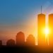 A stylized illustration of the Twin Towers rising above the New York City skyline with a faded American flag in the background at sunrise.