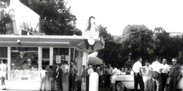 A vintage black and white photo shows customers lined up outside a busy Dari Delite ice cream stand with classic cars parked nearby.