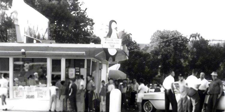 A vintage black and white photo shows customers lined up outside a busy Dari Delite ice cream stand with classic cars parked nearby.