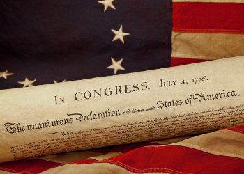 A rolled copy of the Declaration of Independence rests on an American flag with the date July 4, 1776, visible at the top.