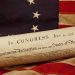 A rolled copy of the Declaration of Independence rests on an American flag with the date July 4, 1776, visible at the top.