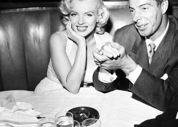 Vintage black and white photo of Marilyn Monroe and Joe DiMaggio sitting together in a restaurant booth and smiling at the camera.