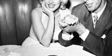 Vintage black and white photo of Marilyn Monroe and Joe DiMaggio sitting together in a restaurant booth and smiling at the camera.