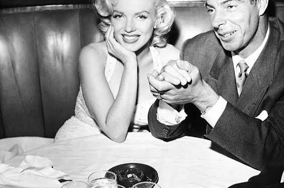 Vintage black and white photo of Marilyn Monroe and Joe DiMaggio sitting together in a restaurant booth and smiling at the camera.