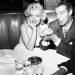 Vintage black and white photo of Marilyn Monroe and Joe DiMaggio sitting together in a restaurant booth and smiling at the camera.