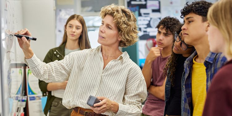 A teacher writes on a whiteboard while several high school students watch during a classroom lesson.