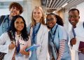 Five smiling medical students in white coats and scrubs pose together while holding notebooks and wearing stethoscopes.