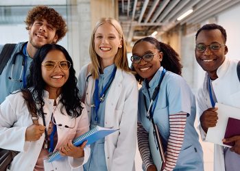 Five smiling medical students in white coats and scrubs pose together while holding notebooks and wearing stethoscopes.