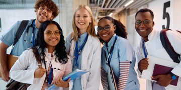 Five smiling medical students in white coats and scrubs pose together while holding notebooks and wearing stethoscopes.