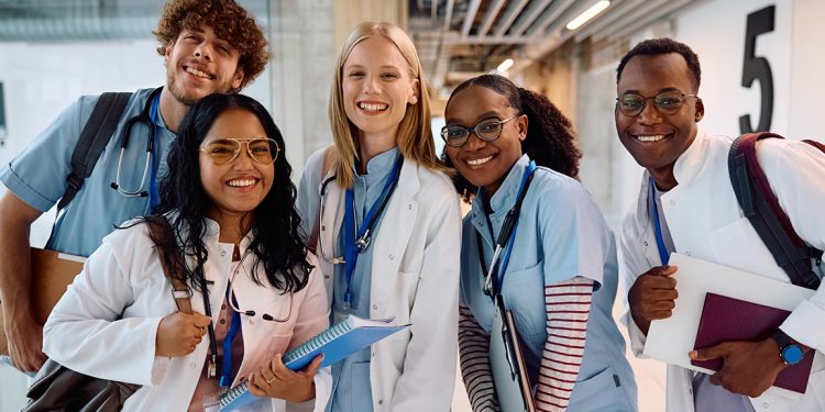 Five smiling medical students in white coats and scrubs pose together while holding notebooks and wearing stethoscopes.