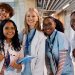 Five smiling medical students in white coats and scrubs pose together while holding notebooks and wearing stethoscopes.