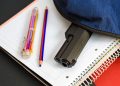 A handgun partially visible inside an unzipped backpack rests on top of school notebooks, illustrating a school safety concern.