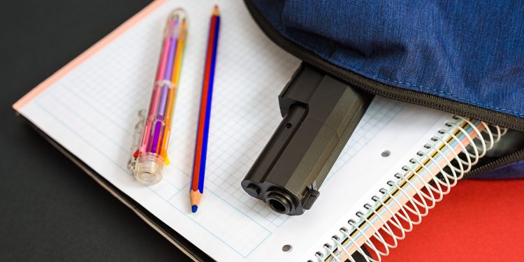 A handgun partially visible inside an unzipped backpack rests on top of school notebooks, illustrating a school safety concern.