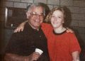 An older man and a young woman smile with their arms around each other while posing indoors in front of a brick wall.