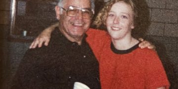 An older man and a young woman smile with their arms around each other while posing indoors in front of a brick wall.