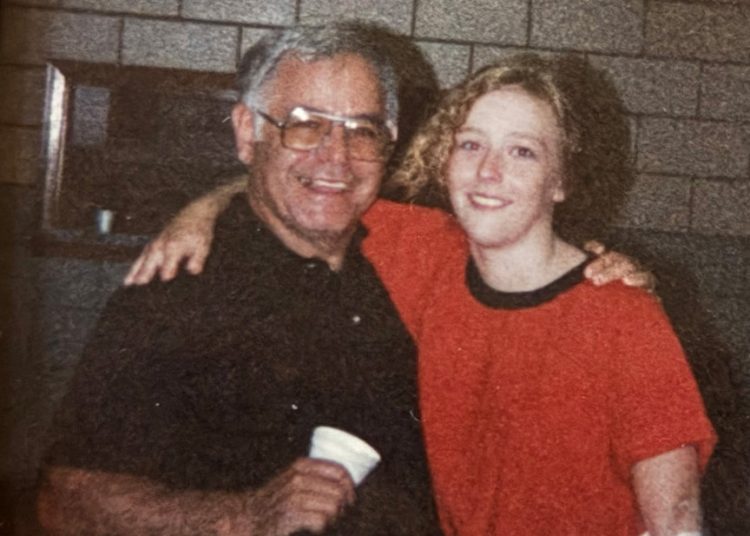 An older man and a young woman smile with their arms around each other while posing indoors in front of a brick wall.