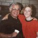 An older man and a young woman smile with their arms around each other while posing indoors in front of a brick wall.