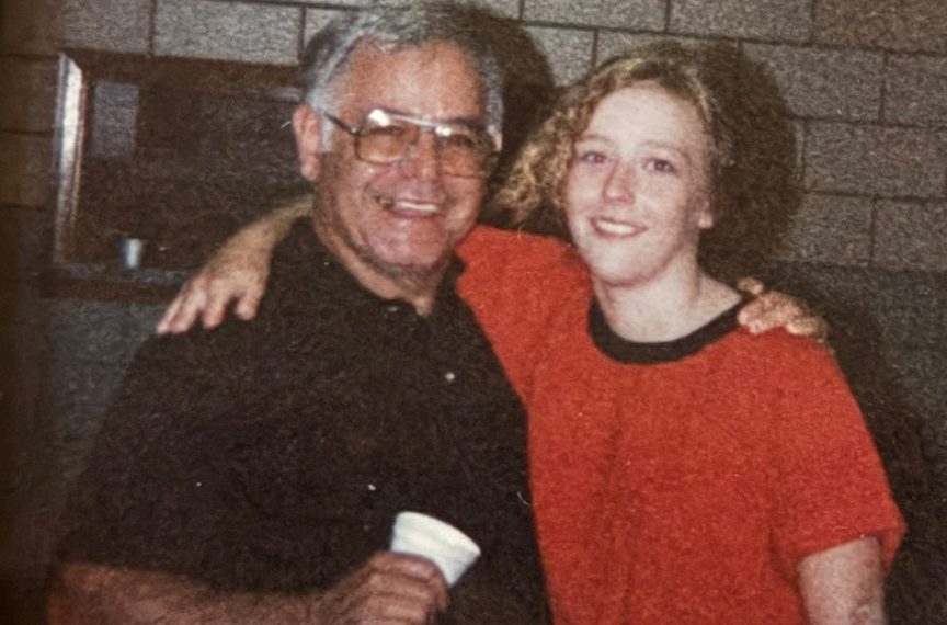 An older man and a young woman smile with their arms around each other while posing indoors in front of a brick wall.