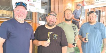 Four men smile while holding trophy awards inside a wood paneled venue following a sporting clays competition.