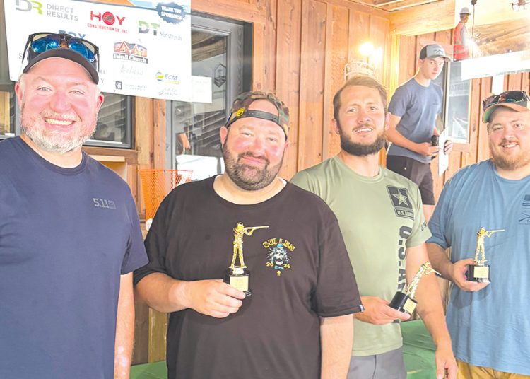 Four men smile while holding trophy awards inside a wood paneled venue following a sporting clays competition.
