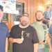 Four men smile while holding trophy awards inside a wood paneled venue following a sporting clays competition.
