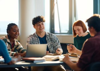 College students collaborate during a group study session around a table with books, notebooks, and a laptop in a bright classroom.