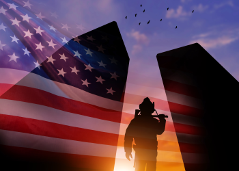 Silhouetted soldier in a spotlight in front of an American flag at sunset with birds in the sky.
