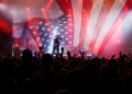 A concert crowd raises their hands beneath a large American flag displayed across a brightly lit stage.