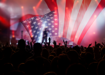 A concert crowd raises their hands beneath a large American flag displayed across a brightly lit stage.