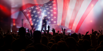 A concert crowd raises their hands beneath a large American flag displayed across a brightly lit stage.