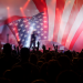 A concert crowd raises their hands beneath a large American flag displayed across a brightly lit stage.