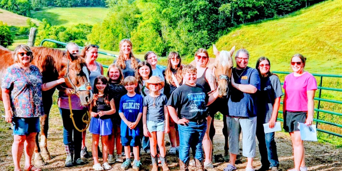Group of adults and children posing beside two horses in a sunny green countryside.