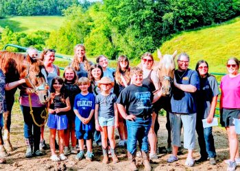 Group of adults and children posing beside two horses in a sunny green countryside.