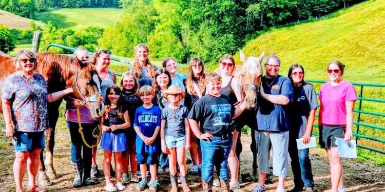 Group of adults and children posing beside two horses in a sunny green countryside.