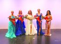 Five women in formal gowns pose on a stage, with the crowned woman in the center holding a bouquet and surrounded by four other women holding flowers.