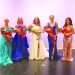 Five women in formal gowns pose on a stage, with the crowned woman in the center holding a bouquet and surrounded by four other women holding flowers.