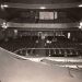 Historic theater auditorium viewed from the stage, with ornate balconies, rows of seats, a central aisle, several people, and a large draped curtain in the foreground.