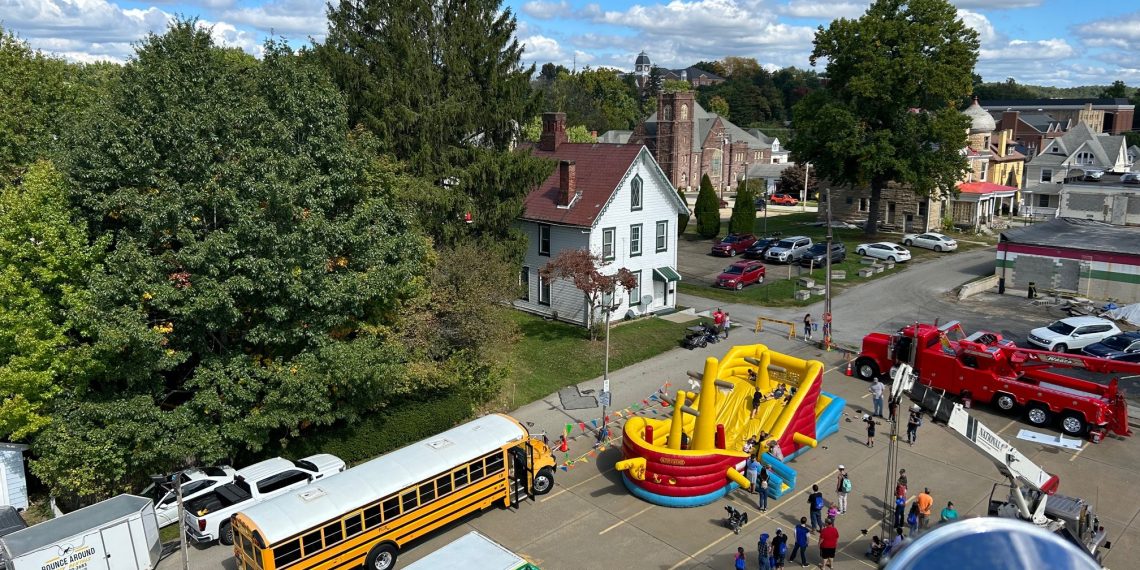 Aerial view of families enjoying a community event with an inflatable obstacle course, school bus, food truck, and emergency vehicles.