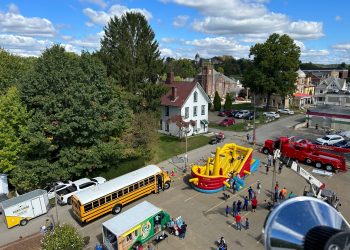 Aerial view of families enjoying a community event with an inflatable obstacle course, school bus, food truck, and emergency vehicles.