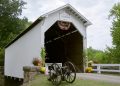 White covered bridge with a clearance sign, a cannon in front, and flowers along the entrance.