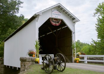 White covered bridge with a clearance sign, a cannon in front, and flowers along the entrance.