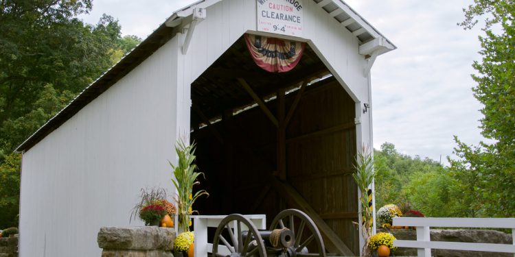 White covered bridge with a clearance sign, a cannon in front, and flowers along the entrance.