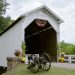 White covered bridge with a clearance sign, a cannon in front, and flowers along the entrance.
