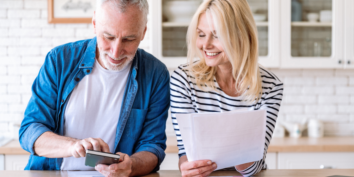 Older couple smiling while reviewing documents at a kitchen counter, with a smartphone and laptop nearby.