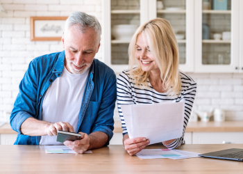 Older couple smiling while reviewing documents at a kitchen counter, with a smartphone and laptop nearby.